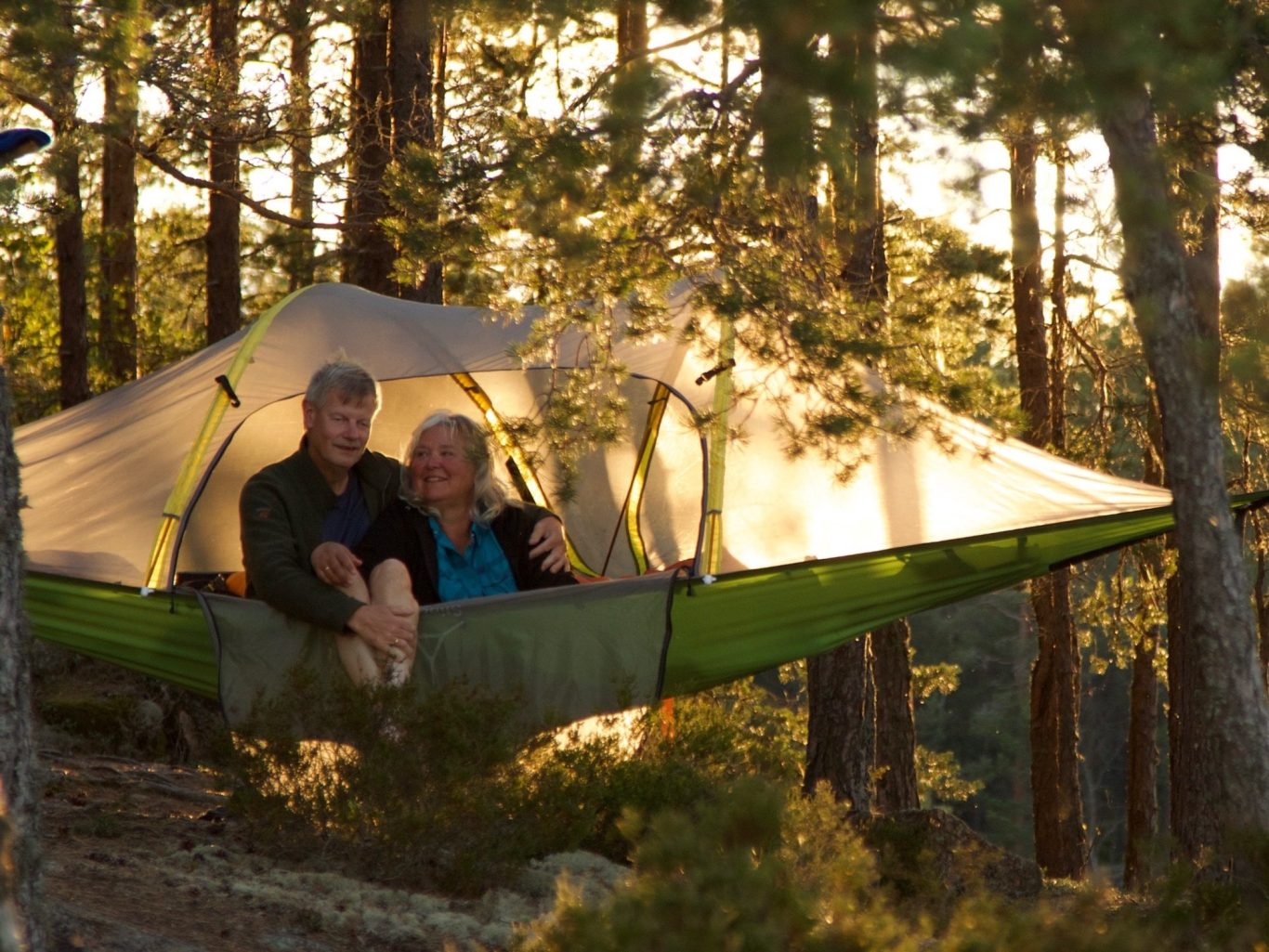 Två personer sitter i ett trädtält mellan träd, omgivna av natur i kvällsljus.