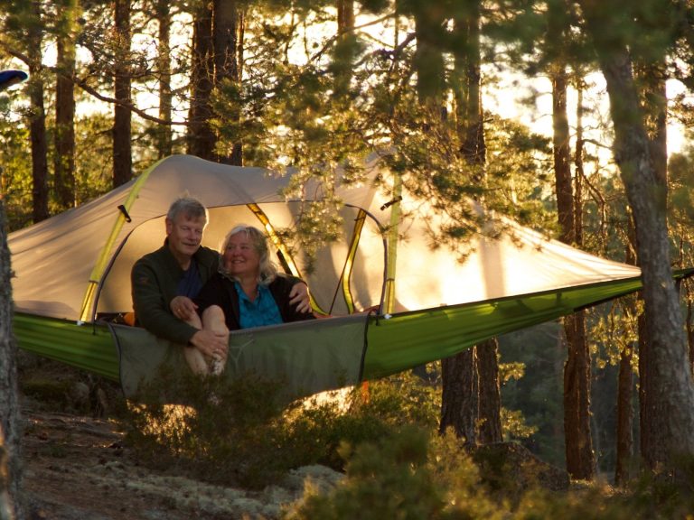Vakna i Tiveden Två personer sitter i ett trädtält mellan träd, omgivna av natur i kvällsljus.