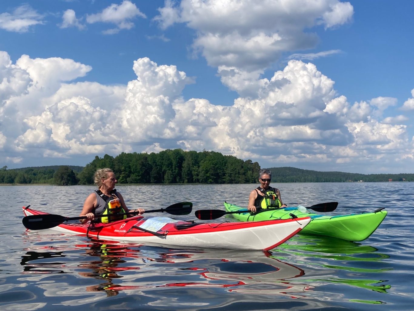 Två personer paddlar kajak på en sjö med gröna omgivningar och molnig himmel.